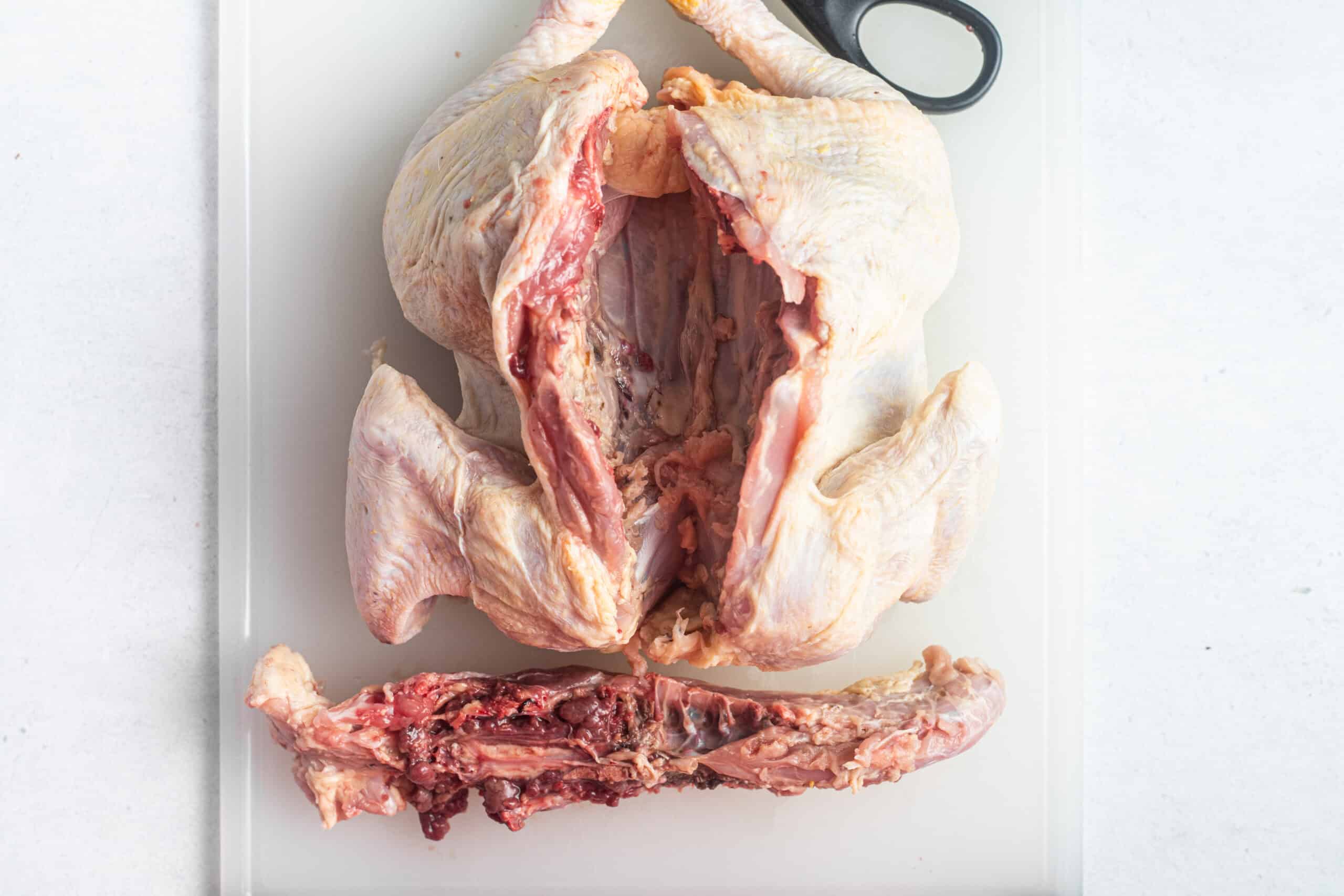 A cutting board with a spatchcocked chicken, a pair of scissors, and a backbone.